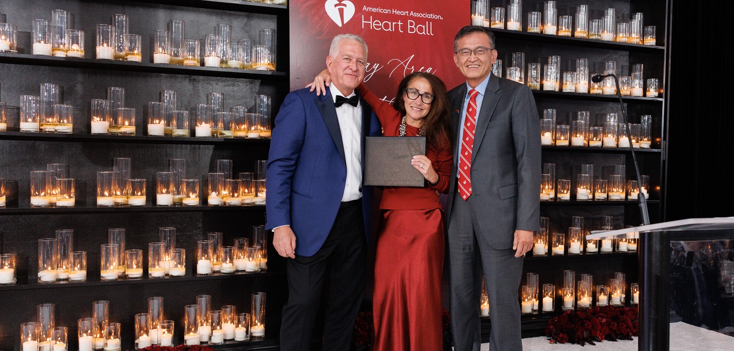 Two men and a woman standing in front of a wall of lit candles and the American Heart Association Heart Ball logo backdrop.